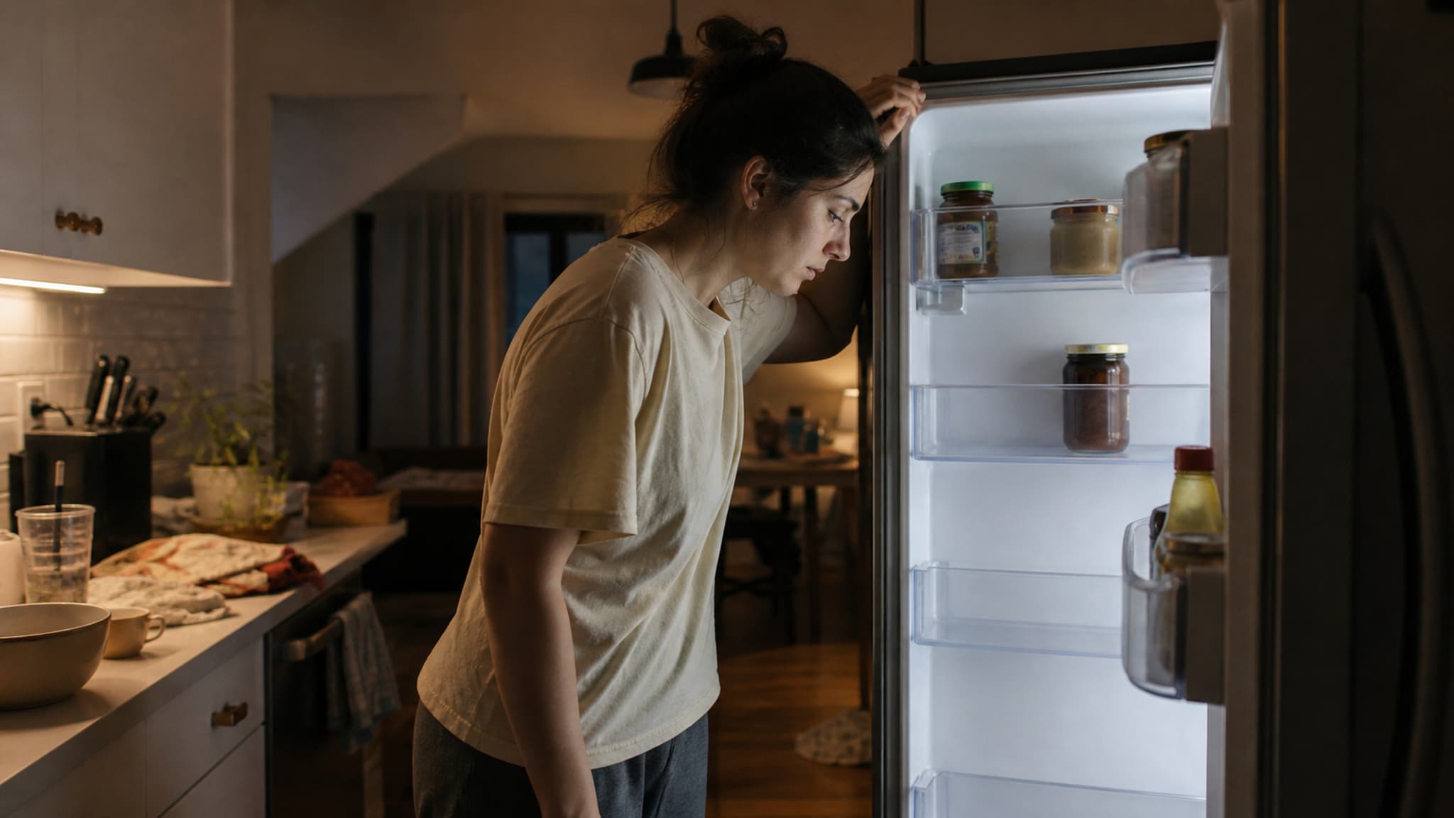 A tired person standing in a kitchen after work, staring at limited meal options and facing the default path of convenience food